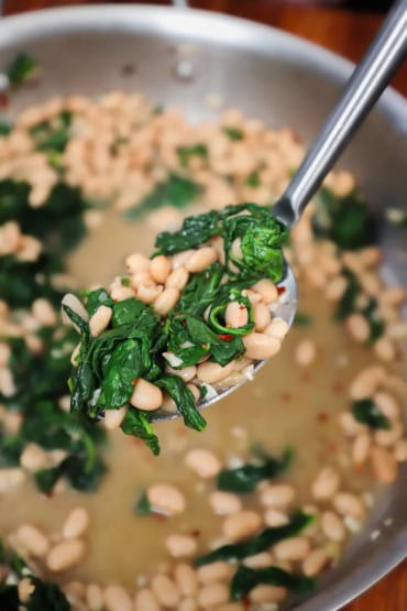 A straight-on view of a serving spoon filled with sautéed white beans and spinach being raised over a skillet filled with the same.