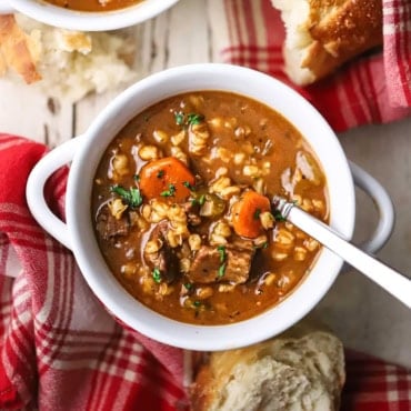 A straight-on view of a large white soup bowl filled with a serving of slow cooker beef and barley soup with chunks of beef, sliced carrots, and barley visible.