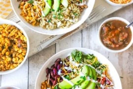 An overhead view of two white bowls that are both filled with a serving of a turkey burrito bowl and they are surrounded by smaller bowls of salsa, pico de gallos, cheese, and corn salsa.