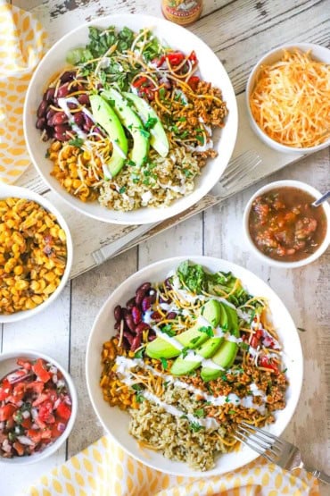 An overhead view of two white bowls that are both filled with a serving of a turkey burrito bowl and they are surrounded by smaller bowls of salsa, pico de gallos, cheese, and corn salsa.