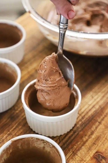 A person using a silver spoon to transfer a chocolate cake batter into a small ramekin that has been greased and dusted with cocoa powder.