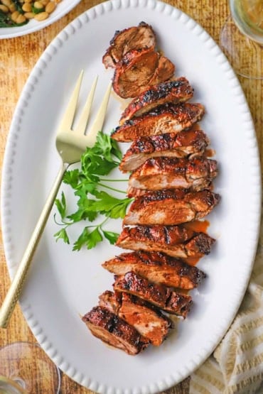 An overhead view of sliced best-ever pork tenderloin topped with a cranberry reduction on a large white platter with sprigs of parsley and a large serving fork next to the sliced meat.