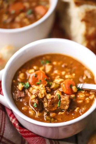 A straight-on view of a large white soup bowl filled with a serving of slow cooker beef and barley soup with chunks of beef, sliced carrots, and barley visible.