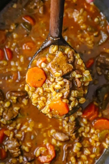 An overhead view of a wooden ladle lifting up a helping of beef and barley soup form a slow cooker filled with the same.