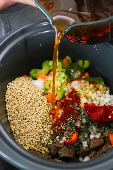 A person pouring beef stock from a large glass measuring cup into a slow cooker filled with uncooked barley, sliced vegetables, seared beef chunks, hers, and tomatoes.