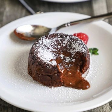 A chocolate lave cake sitting on a white dessert plate that has been dusted with powdered sugar with a fresh cranberry sitting next to it.