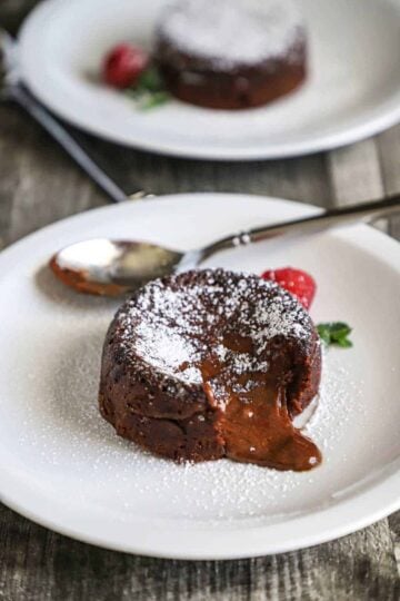 A chocolate lave cake sitting on a white dessert plate that has been dusted with powdered sugar with a fresh cranberry sitting next to it.