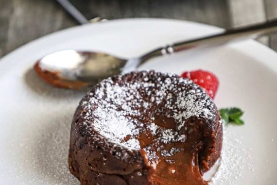 A chocolate lave cake sitting on a white dessert plate that has been dusted with powdered sugar with a fresh cranberry sitting next to it.