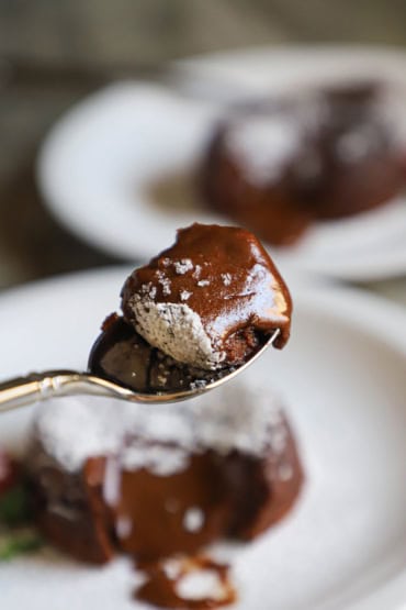 A spoon being raised with a serving of gooey chocolate lava cake on it with the half-eaten cake in the background.