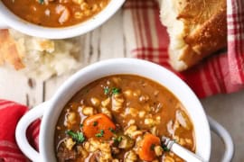 An overhead view of two white soup bowls filled with slow cooker beef and barley soup with red checker napkins and a piece of bread nearby.