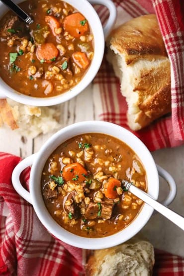 An overhead view of two white soup bowls filled with slow cooker beef and barley soup with red checker napkins and a piece of bread nearby.