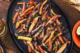 An overhead view of a large fajita skillet that is filled with a serving of veggie fajitas and surround by festive bowl of cheese, limes, sour cream, and a stack of charred flour tortillas.