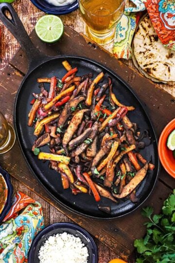 An overhead view of a large fajita skillet that is filled with a serving of veggie fajitas and surround by festive bowl of cheese, limes, sour cream, and a stack of charred flour tortillas.