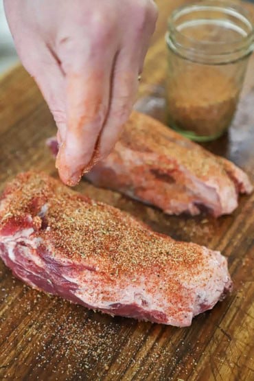 A person sprinkling a pork rub onto two uncooked pork wings that are resting on a wooden cutting board next to a jar half-filled with the rub.