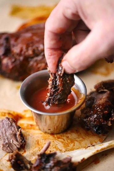 A straight-on view of a person plunging a piece of BBQ pork wings into a small silver vessel filled with homemade barbecue sauce.