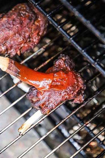 A close-up view of a person using a small kitchen brush to apply barbecue sauce onto a pork wing that is on a metal grate of an electric smoker.