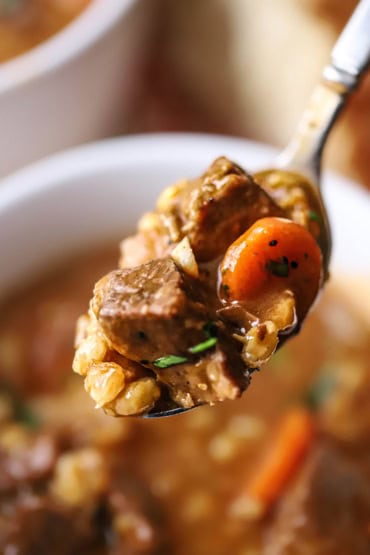 A close-up view of a silver spoon holding up a serving of slow cooker beef and barley soup with beef chunks, sliced carrot, and barley visible.