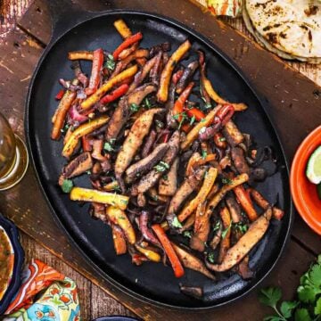 An overhead view of a large fajita skillet that is filled with a serving of veggie fajitas and surround by festive bowl of cheese, limes, sour cream, and a stack of charred flour tortillas.