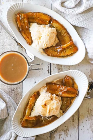 An overhead view of two oval dessert dishes that are filled with bananas foster and topped with a scoop of melting vanilla ice cream and a mug of creamed coffee nearby.