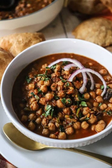 A straight-on view of a bowl of homemade chana masala toped with chopped cilantro and two slivers of red onion with homemade poori bread nearby.