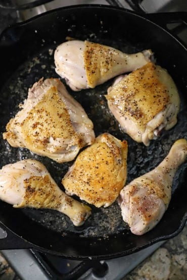 An overhead view of a bone-in chicken legs and chicken thighs with skin that have been seared and browned in a large black cast-iron skillet in lard.