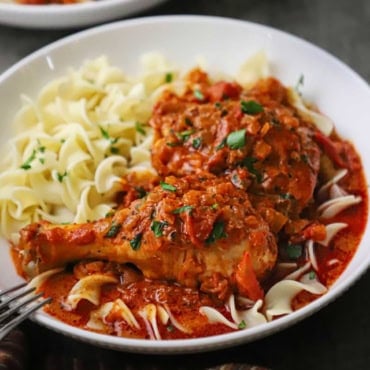 A close-up view of a shallow white bowl that is filled with a serving of chicken paprikash and cooked egg noodles garnished with chopped Italian parsley.