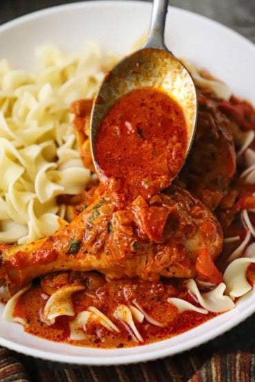 A close-up view of a person using a serving spoon to transfer chicken paprikash sauce over a cooked chicken leg in a shallow bowl also filled with cooked egg noodles.