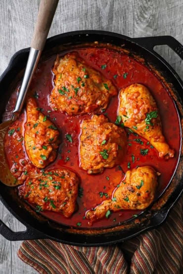 An overhead view of a large black cast-iron skillet filled with chicken paprikash including a vibrant red sauce and browned chicken pieces.