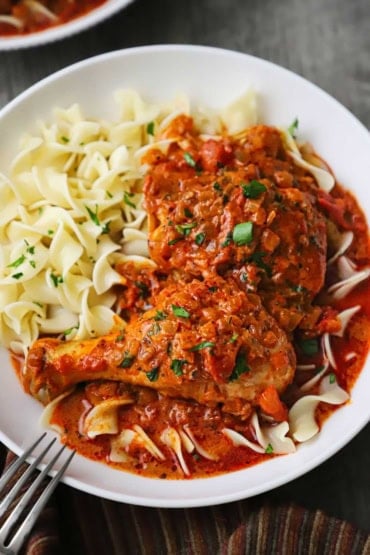 An overhead view of a white shallow bowl holding a serving of chicken paprikash which included two pieces of cooked chicken and topped with a bright red paprika sauce with tomatoes.