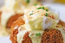 A close-up view of an untouched crab cakes egg Benedict with hollandaise flowing over the top of the poached egg and pooling on a white plate.