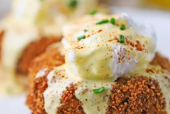 A close-up view of an untouched crab cakes egg Benedict with hollandaise flowing over the top of the poached egg and pooling on a white plate.