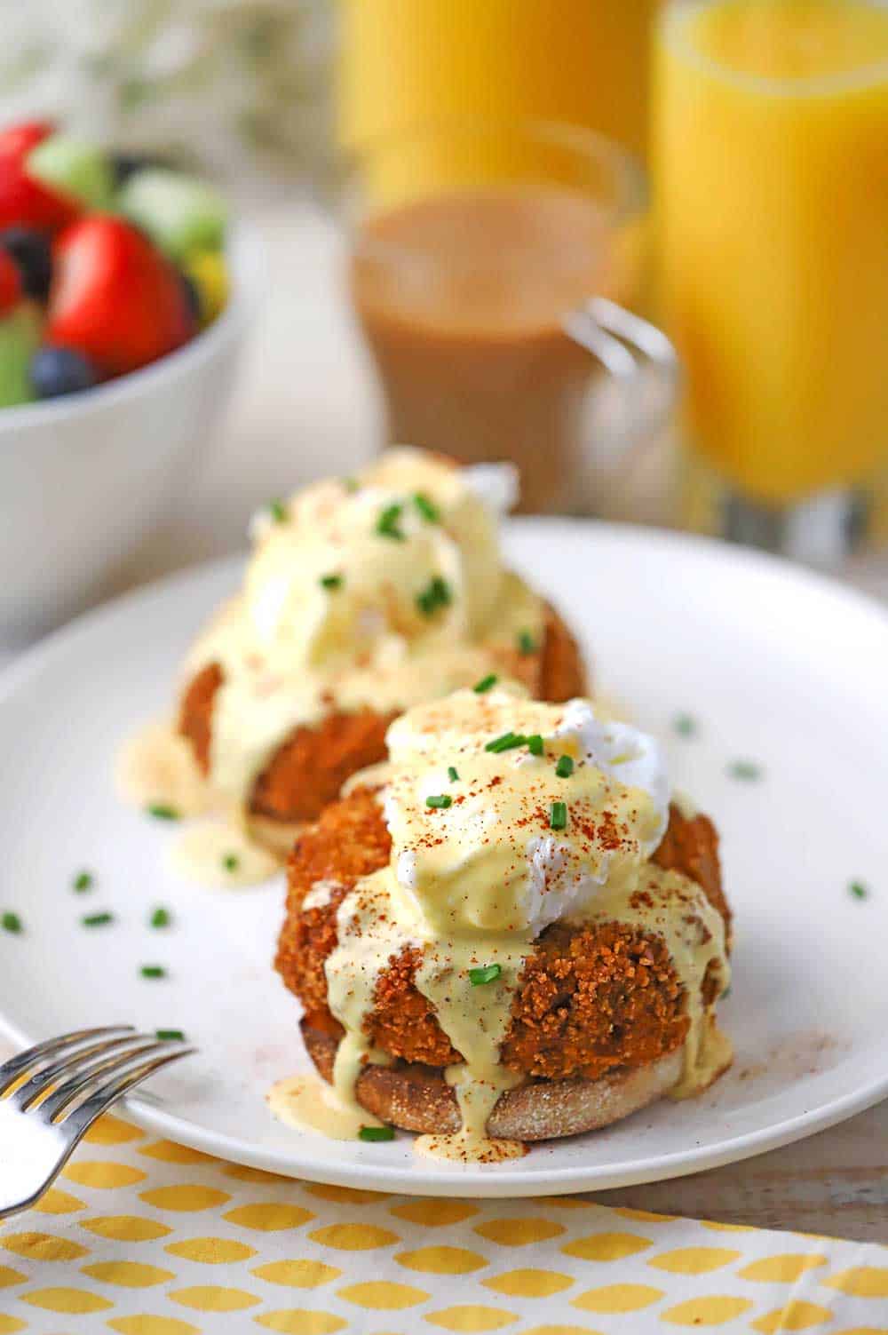 A straight-on view of two crab cake Benedict servings on a single white dinner plate with a tall glass of orange juice and a bowl of fruit in the background