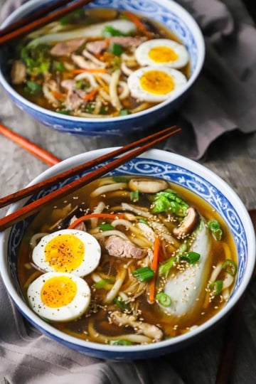 A straight-on view of two colorful Asian soup bowls filled with servings of udon soup with pork topped with hard boiled eggs and chopped scallions.