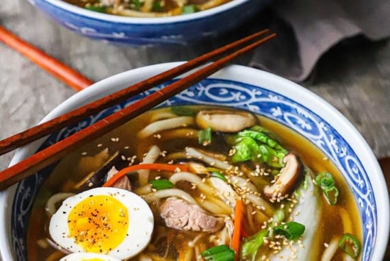 A straight-on view of two colorful Asian soup bowls filled with servings of udon soup with pork topped with hard boiled eggs and chopped scallions.