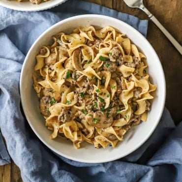 An overhead view of a white bowl filled with a serving of weeknight beef stroganoff with a blue napkin wrapped around the base of the bowl.