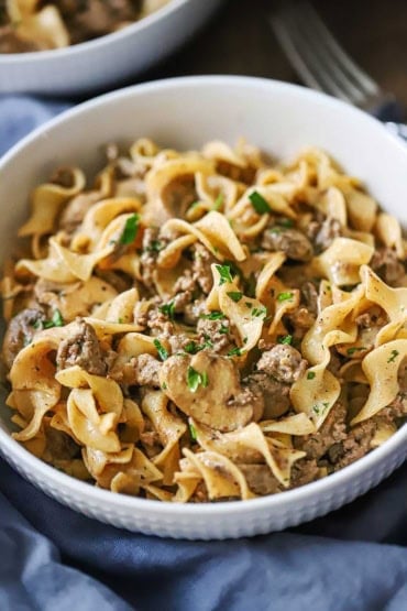 A close-up view of a white shallow bowl filled with a serving of weeknight beef stroganoff with a light blue napkin wrapped around the bottom of the bowl.