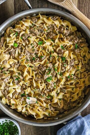 An overhead view of a large straight-edge stainless steel skillet filled with weeknight stroganoff of a creamy ground beef sauce tossed with egg noodles.