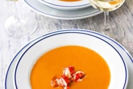 A straight-on view of two white shallow bowl filled with servings of lobster bisque with two glasses of white wine near by and chunks of fresh lobster on top of the soups.