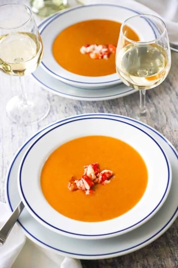 A straight-on view of two white shallow bowl filled with servings of lobster bisque with two glasses of white wine near by and chunks of fresh lobster on top of the soups.