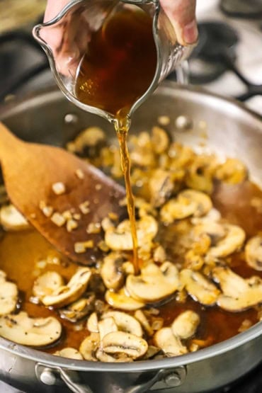 A person pouring beef broth from a glass measuring cup into a large silver skillet filled with sautéed sliced mushrooms.