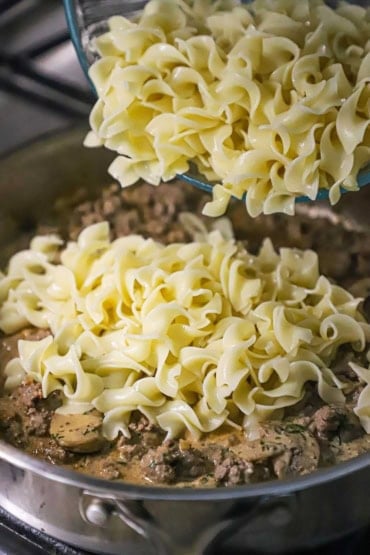 A close-up view of a cooked egg noodles being transferred from a glass bow into a skillet filled with a creamy sauce, ground beef, and sautéed mushrooms.