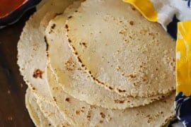 A side view of a stack of homemade corn tortillas that are resting on a wooden board topped with a colorful napkin with a bowl of salsa and soften butter nearby.