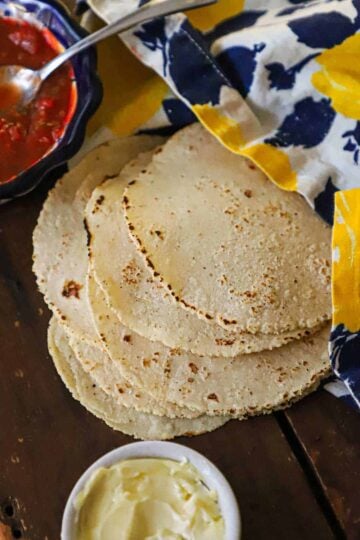 A side view of a stack of homemade corn tortillas that are resting on a wooden board topped with a colorful napkin with a bowl of salsa and soften butter nearby.