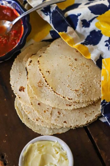 A side view of a stack of homemade corn tortillas that are resting on a wooden board topped with a colorful napkin with a bowl of salsa and soften butter nearby.