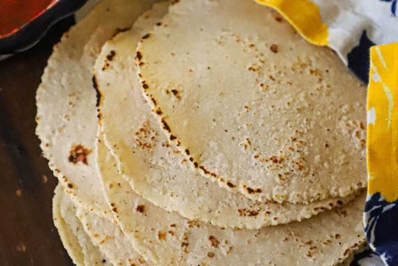 A side view of a stack of homemade corn tortillas that are resting on a wooden board topped with a colorful napkin with a bowl of salsa and soften butter nearby.