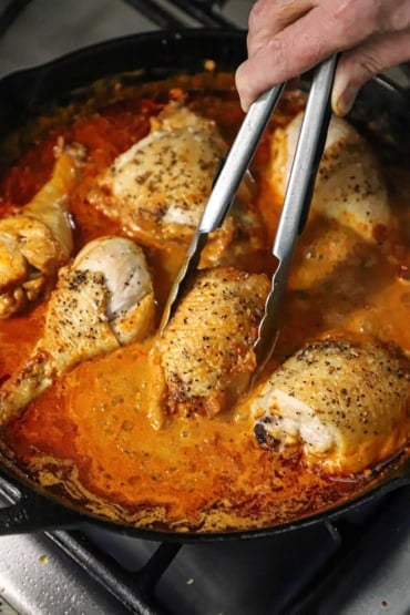 A person using a pair of metal tongs to nestle a seared chicken thigh into a large skillet of a bright red simmering sauce with other chicken pieces in the sauce, as well.