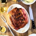 A large Berkshire ham roast that has been scored on top resting on a white oval platter and surrounded by various side dishes.