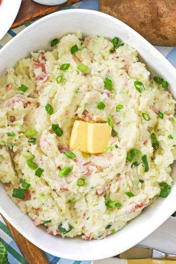 An overhead view of a large bowl filled colcannon (Irish mashed potatoes) with a couple tabs of butter in the center and whole russet potatoes resting nearby.