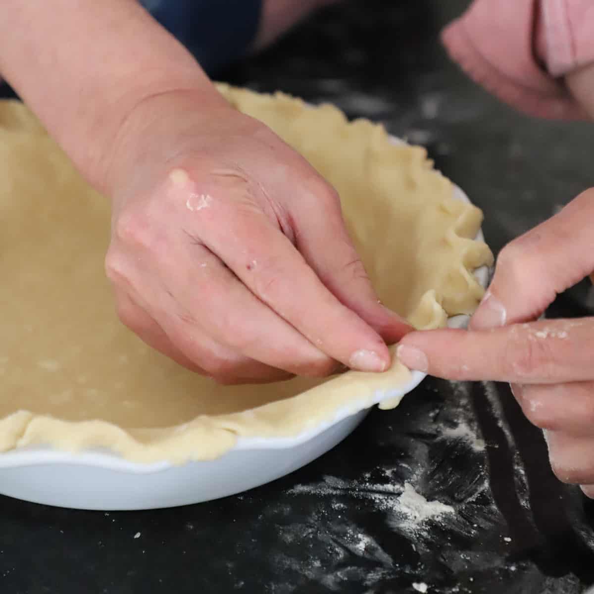 A person using his two index fingers to flute the top of an uncooked pie dough that has been fitted into a pie dish. 