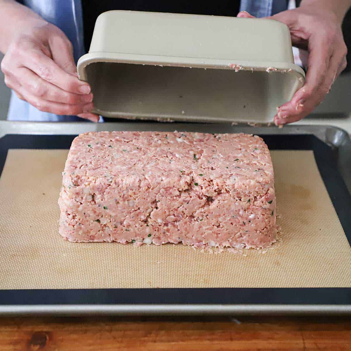 A person pulling a loaf pan away from an inverted uncooked ham loaf on a silicone mat-lined baking sheet. 